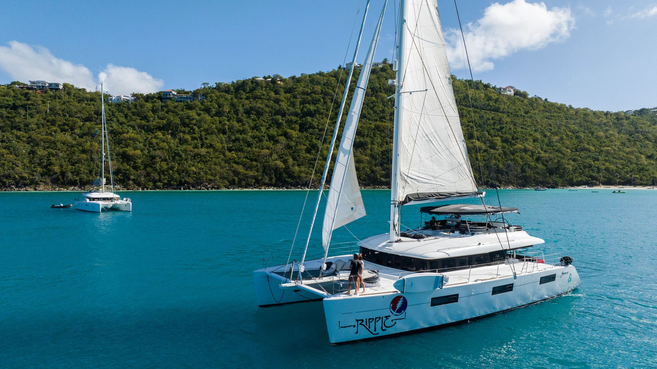 Ripple catamaran sailing with guests on deck near forested hillside